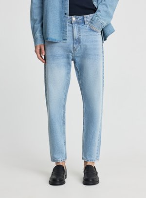 Persona che indossa jeans chiari a gamba stretta, una camicia di jeans coordinata, mocassini neri e una maglia scura, in piedi con una mano in tasca.