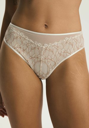 Kobieta w beżowej koronkowej bieliźnie z kwiatowymi wzorami i półprzezroczystymi panelami z siateczki, pokazująca środkową część ciała i górne partie ud.