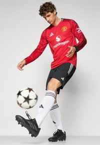 Piros Manchester United mez fekete csíkokkal, fekete sorttal, fekete cipővel és fehér zoknival. Egy fehér csillagmintás focilabda található mellette.