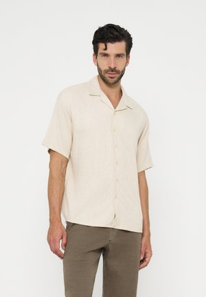 Homme aux cheveux foncés et à la barbe portant une chemise beige à manches courtes boutonnée et un pantalon vert olive, debout devant un fond blanc uni.