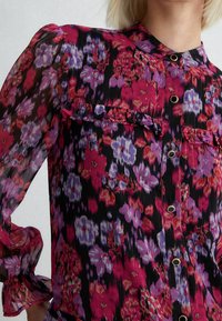 Blusa floreale nera, con motivi rosa e viola, dettagli arricciati sulle tasche e accenti di bottoni dorati. Tessuto trasparente con maniche a sbuffo.