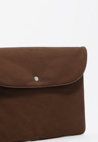 Brązowa torba w stylu koperty, wykonana z gładkiego materiału, z zaokrągloną klapą zamykaną na srebrny nap oraz eleganckim, minimalistycznym wzornictwem.