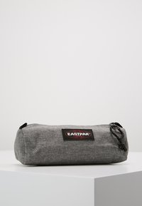 Gray Eastpak pencil case, cylindrical shape, made of textured fabric, features a black logo patch, zip closure, and black drawstring.