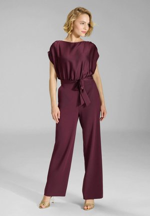 Femme debout en haut en satin bordeaux et pantalon large assorti avec ceinture à la taille, portant des talons dorés, regardant sur le côté sur fond gris.