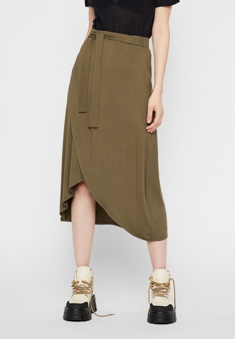 Personne portant une jupe portefeuille vert olive avec une ceinture, un haut noir et de grosses bottines beiges à lacets, debout devant un fond uni.