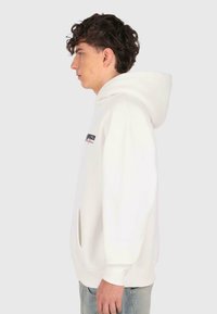 Ung mand med krøllet hår iført en hvid hættetrøje og lyseblå jeans, stående i profil mod en ensfarvet lys baggrund.