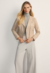 Beżowa skórzana kurtka motocyklowa z asymetrycznym zamkiem błyskawicznym i bocznymi kieszeniami, w połączeniu z szarym golfem i jasnoszarymi spodniami.