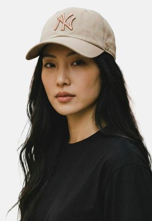 Frau mit langen schwarzen Haaren trägt eine beige NY-Baseballkappe und ein schwarzes Shirt, blickt geradeaus mit neutralem Gesichtsausdruck vor einem schlichten hellen Hintergrund.