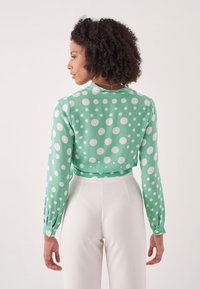 Grüne Bluse mit Punkten, langen Ärmeln und geknöpften Manschetten; leichtes Material, cropped Design. Getragen mit hochgeschnittenen weißen Hosen.