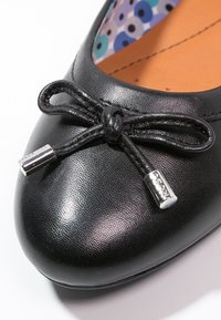 Gros plan d'une ballerine en cuir noir avec un nœud décoratif et des embouts métalliques argentés sur la zone des orteils.