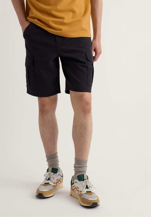 Uomo che indossa pantaloncini cargo neri, maglietta giallo senape, calzini beige e scarpe da ginnastica bianche con accenti verdi e arancioni, in piedi davanti a uno sfondo bianco.