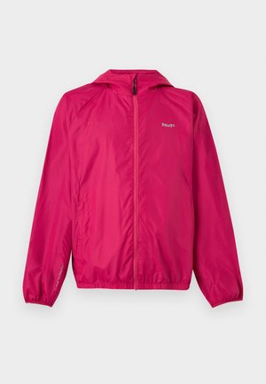 Jeune homme portant une veste à capuche zippée rose vif et un jean gris clair, debout devant un fond blanc uni.
