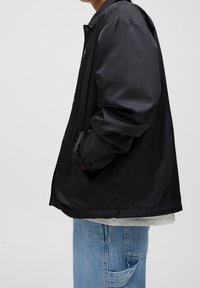 Czarna nylonowa kurtka o luźnym kroju, z elastycznymi mankietami, bocznymi kieszeniami i zapięciem na zamek, zestawiona z jasnoniebieskimi dżinsami.