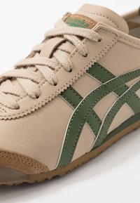 Beige lædersneakers med grønne accenter, der har teksturerede snørebånd og syninger samt en brun gummisål. Bemærkelsesværdig branding på tungen.