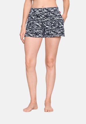 Femme portant un short imprimé zèbre noir et blanc, les mains dans les poches, debout pieds nus devant un fond uni.