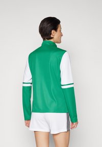 Grøn langærmet sportstrøje med høj krave, hvide ærmer og grønne striber på armene, parret med hvide sportsshorts.