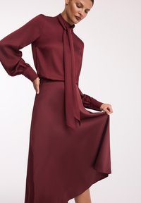 Burgundowa jedwabna sukienka z wysokim kołnierzem, długimi bufiastymi rękawami i wiązaną kokardą, z falującym asymetrycznym dołem.