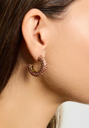Guld øreringe med hoops dekoreret med lyserøde rhinsten, der har et buet design. Nærbillede fremhæver overfladens struktur og glimt.