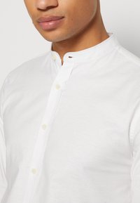 Camisa de algodón blanca con cuello tipo banda, adornada con pequeños botones y una abertura abotonada en el cuello. Textura suave.