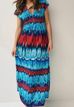 Kobieta w bezrękawną sukienkę maxi z dekoltem w serek, z niebiesko-czerwono-granatowym wzorem typu tie-dye, stojąca na tle jednolitej jasnej ściany.