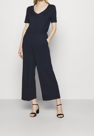 Vrouw draagt een marineblauwe jumpsuit met korte mouwen, V-hals en wijde pijpen, gecombineerd met zwarte sandalen met enkelbandje en blokhak, staand tegen een effen muur.