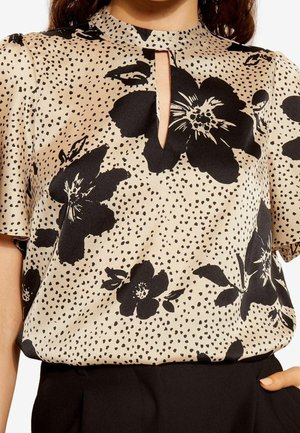Beige bluse med sorte store blomster- og små prikmønstre, korte ærmer og nøglehul-udskæring, stoppet ned i sorte bukser.