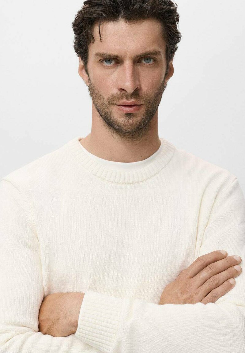 Weißer gestrickter Pullover mit rundem Ausschnitt und gerippten Bündchen. Das Modell hat die Arme verschränkt und zeigt die glatte Textur und Passform des Kleidungsstücks.