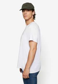 White cotton t-shirt with short sleeves, paired with blue jeans and a dark green baseball cap featuring a front logo detail.