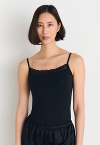 Svart cami-top med tynne stropper og blondekant ved halslinjen. Stoffet ser glatt og tettsittende ut, kombinert med løse svarte bukser.