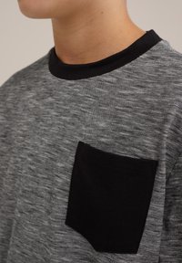 Siva in črna črtasta majica s črnim žepom in črnimi rebrastimi izrezom; tkanina se zdi mehka in fino strukturirana.