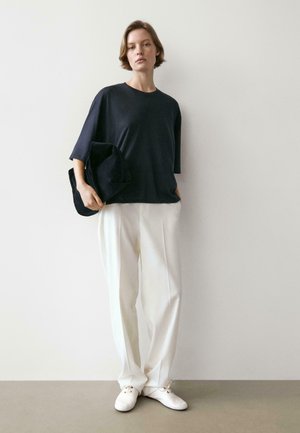 Frau steht an einer Wand, trägt ein weites schwarzes Oberteil, weiße, weit geschnittene Hosen, weiße Schuhe und hält eine schwarze Clutch.