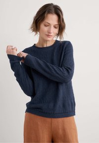 Navy textured sweater with a ribbed neckline and cuffs, featuring a patterned weave. Paired with light brown linen pants for contrast.