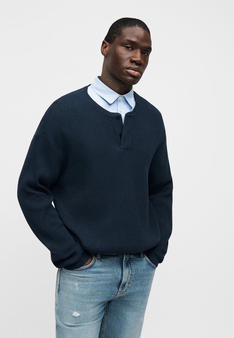 Pull texturé bleu marine avec col rond, patte de boutonnage et manches longues, superposé à une chemise à col bleu clair, porté avec un jean.