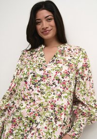 Bloes met bloemenprint in pastelkleuren van roze, groen en zwart. Heeft een V-hals en bolle mouwen met gathered manchetten, gemaakt van lichtgewicht stof.