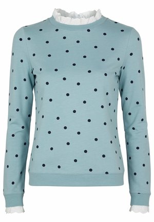 Hellblaues Langarmhemd mit unregelmäßigen schwarzen Polka-Dots, weißer Rüschenspitze am Ausschnitt und an den Manschetten.