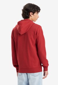 Felpa rossa con cappuccio e coulisse, polsini e orlo a coste. Il tessuto appare morbido, con una texture liscia e senza motivi visibili.