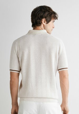 Mand med kort brunt hår set bagfra, iført en hvid tekstureret kortærmet strikket poloshirt med brune kanter ved manchetterne og hvide bukser.