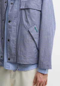 Μπλε και λευκό καρό μπουφάν με κουμπιά σκρατς, τσέπη με καπάκι, φορεμένο πάνω από ανοιχτό μπλε πουκάμισο και μπεζ παντελόνι, εμφανίζοντας το χέρι ενός άνδρα.