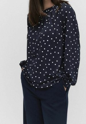 Kvinde iført en mørkeblå bluse med hvide prikker og mørkeblå bukser, hænderne i lommerne, foran en ensartet lys baggrund.