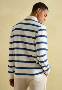 Koszulka polo z długim rękawem wykonana jest z białej tkaniny w poziome granatowe paski, ma klasyczny kołnierzyk oraz boczne rozcięcia u dołu, co umożliwia swobodę ruchów.