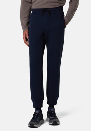 Uomo che indossa pantaloni da jogging blu navy con coulisse in vita, abbinati a sneaker nere e grigie e una maglia grigia a maniche lunghe.
