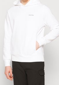 Witte hoodie met een voorzak, gemaakt van zachte stof. Bevat een klein grijs logo op de linkerborst en ribgebreide manchetten en onderrand.