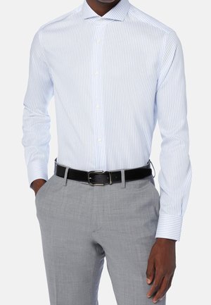 Mann trägt hellblau-weiß gestreiftes langärmliges Hemd, graue Anzughose und einen schwarzen Ledergürtel, Hand in der Tasche.