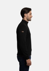 Jersey negro de punto con un alto cuello, puños acanalados y una pequeña etiqueta de cuero marrón en el hombro. Se muestra en vista lateral.