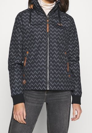Person, der bærer en sort lynlåstrøje med et gråt zigzagmønster, læder lynlås træk og sorte ribmanchetter, kombineret med mørkegrå jeans.