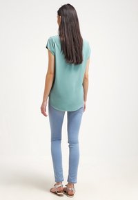 Ljusgrön kortärmad blus med rundad nederkant, istället ihop med ljusblå smala jeans och sandaler med silverdekorationer.