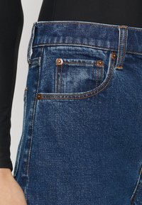 Jeans in denim blu scuro con taglio classico, design a cinque tasche, rivetti in rame e dettagli di cucitura arancio a contrasto.