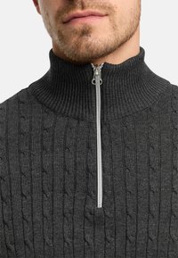 Mörkgrå zippered tröja med hög krage och texturerat kabelstickat mönster, med en silverfärgad dragkedja som accent.