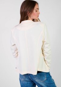 Veste cintrée en crème clair, en matériau lisse, avec des épaules structurées, des manches longues et des détails sur les coutures latérales.