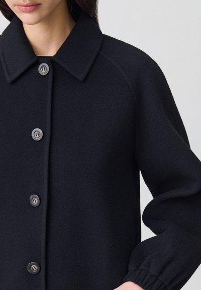 Veste en laine noire avec un col, une fermeture à boutons et des poignets froncés. Texture lisse avec un subtil éclat et quatre boutons visibles à l'avant.